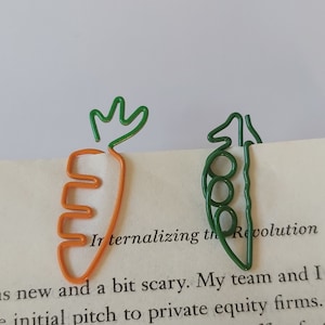 May include: Two wire bookmarks shaped like a carrot and a pea pod. The carrot is orange and the pea pod is green. The bookmarks are on a book with the text "Internalizing the Revolution | 7" visible.