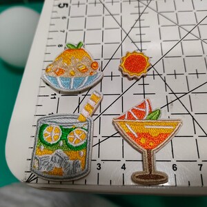 May include: Four embroidered iron-on patches featuring summer-themed designs: a glass of lemonade with a straw, a slice of orange in a martini glass, a scoop of orange sherbet, and a simple orange sun.