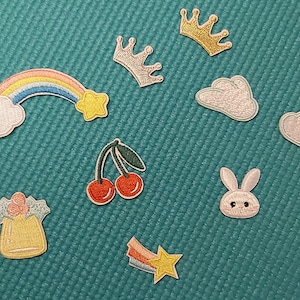 Puede incluir: Un conjunto de diez parches para planchar con varios diseños lindos, que incluyen un arcoíris, nubes, un conejito, cerezas, una estrella y coronas. Los parches son todos blancos, rosas, amarillos, azules y verdes.