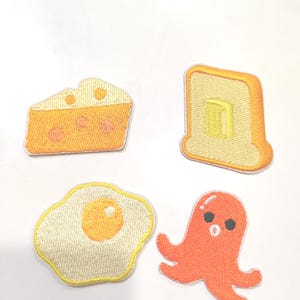 Peut inclure: Ensemble de quatre écussons brodés sur le thème de la nourriture. Comprend une part de fromage, une tranche de pain grillé beurrée, un œuf au plat et une pieuvre orange. Chaque écusson est finement brodé avec des couleurs vives.
