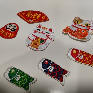 Puede incluir: Un conjunto de siete parches bordados para planchar con motivos de amuletos y símbolos japoneses de la buena suerte. Los parches incluyen una muñeca daruma roja y dorada, un maneki-neko (gato de la suerte) blanco y dorado, un abanico rojo y dorado con el texto "有銭" (que significa "dinero"), un pez koi rojo y dorado, un pez koi azul y rojo, un pez koi verde y dorado y un gato blanco y naranja que sostiene un pez koi rojo y dorado.