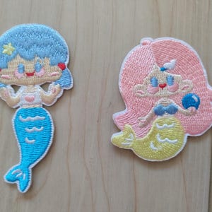 Puede incluir: Dos parches bordados de sirenas. Uno tiene cabello azul y una cola turquesa. El otro tiene cabello rosa y una cola amarilla, sosteniendo una bola azul. Ambos tienen un estilo de dibujos animados con costuras detalladas.
