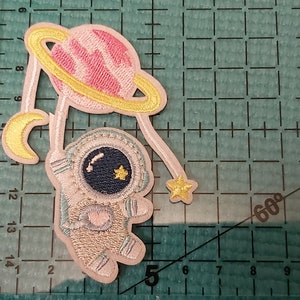 May include: An embroidered patch featuring an astronaut in a white spacesuit holding a pink planet with a yellow ring. The astronaut has a blue helmet with a yellow star on it. The patch is decorated with a yellow crescent moon and a yellow star.