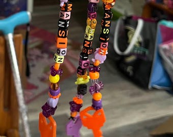 May include: Two beaded keychains with the words "HAUNTED MANSION" spelled out in colorful letter beads. The keychains feature star, flower, and key charms in orange, purple, and yellow. The keychains are held up by a silver ring.