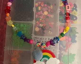 May include: A colorful beaded necklace with a rainbow and cloud charm. The necklace features a variety of beads in rainbow colors, including stars and other shapes. The necklace is displayed in a clear plastic organizer.