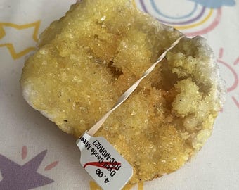 May include: A rough, yellow geode with a tag attached. The geode has a crystalline structure and is a mix of yellow and white. The tag reads "Dyed Geode Mixe" and "Roc-MOR027". The geode is resting on a patterned fabric with stars and rainbows.