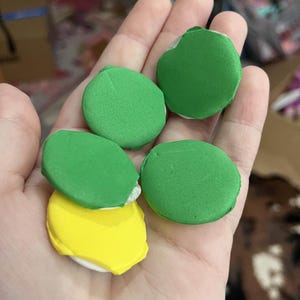 May include: A hand holding five colorful macarons. Four are green, and one is yellow. The macarons are round with a white base. The background is blurred, suggesting an indoor setting.