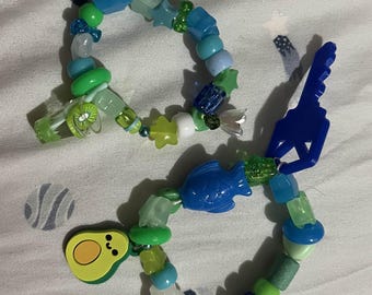 May include: Two beaded bracelets with blue, green, and clear beads. One bracelet features a blue dolphin charm, while the other has a blue fish and avocado charm. The bracelets have various shapes and sizes of beads.