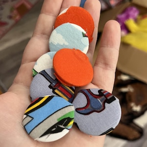 May include: A selection of fabric-covered buttons in various colours and patterns. The buttons feature designs with clouds, geometric shapes, and solid orange. The buttons are round and appear handmade.