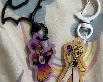 May include: Two acrylic keychains featuring cartoon characters. One keychain has a bat-shaped clasp and a character with a bat-themed costume. The other keychain has a white clasp and a character with a Halloween-themed costume.