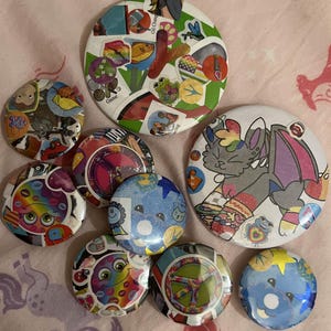 May include: A collection of colorful, round pinback buttons with various designs. Some buttons feature cartoon characters, peace signs, and sticker collages. The buttons are scattered on a pink patterned surface.