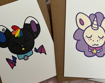 May include: Two greeting cards with cartoon animal illustrations. One card features a black bat with rainbow accents and a blue and purple color scheme. The other card has a unicorn-like creature with a purple mane and a star necklace.
