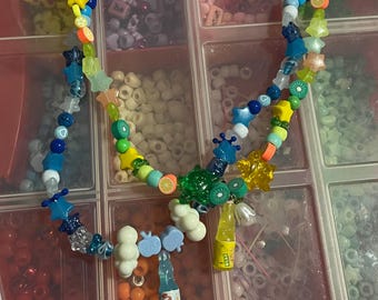 May include: A colorful beaded necklace with various charms, including stars, smiley faces, and fruit slices. The necklace features a mix of blue, yellow, and clear beads, with small bottle and cloud charms. The necklace is displayed in a clear plastic organizer.