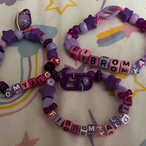 May include: Three handmade beaded bracelets in shades of purple, pink, and white. The bracelets feature star-shaped beads and letter beads spelling out "FIBROMYALGIA" and a name. The bracelets are on a patterned surface.