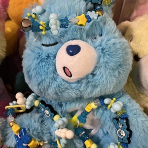 Op de afbeelding: Een blauwe Care Bear knuffel met een gele, blauwe en witte kralenketting en een kroon van blauwe, gele en witte kralen. De Care Bear heeft een blauwe neus en een witte buik. De Care Bear zit op een doos met de tekst "Care Bears" met een hart.