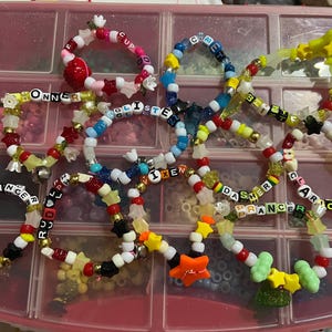 May include: A collection of colorful beaded bracelets with various designs and text. The bracelets feature a mix of red, white, black, yellow, and blue beads, with some spelling out words like "Dancer" and "Prancer".