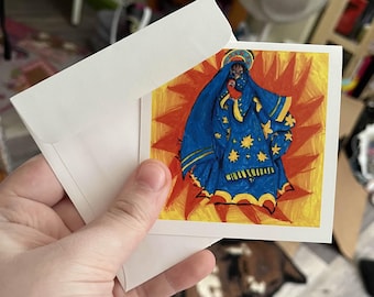 May include: A square art print featuring a religious figure in a blue robe adorned with gold stars, set against a vibrant orange and yellow background. The print is held with a white envelope, suggesting a greeting card or art print.