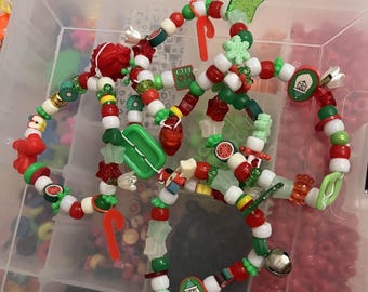 May include: Several beaded bracelets with Christmas-themed charms and beads. The bracelets feature red, green, white, and gold beads, along with charms like candy canes, stars, and Santa Claus faces. The bracelets are arranged in a clear plastic container.