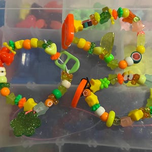 May include: Brightly coloured beaded bracelets with charms such as stars, flowers, and fruit slices. The bracelets use orange, yellow, green, and clear beads. The charms give the jewellery a playful and whimsical appearance.