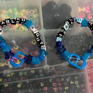 May include: Two beaded bracelets. One bracelet spells "DISABLED RIGHTS" with black and white letter beads. The bracelets are connected by a blue plastic piece. The bracelets have blue star and glitter accents.