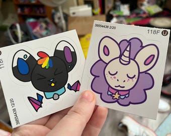 May include: Two stickers featuring cartoon animal designs. One is a black mouse with rainbow hair and a blue collar. The other is a purple unicorn-like creature with a star necklace. Each sticker has a unique serial number.