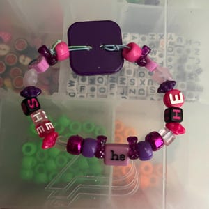 May include: A handmade bracelet with a purple square clasp and a variety of beads. The bracelet features pink, purple, and clear beads, with some spelling out "SHE" and "HE" and the word "he".