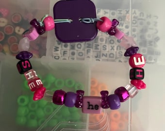 May include: A handmade bracelet with a purple square clasp and a variety of beads. The bracelet features pink, purple, and clear beads, with some spelling out "SHE" and "HE" and the word "he".