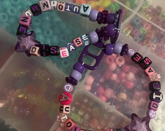 May include: Two handmade beaded bracelets. One bracelet spells out "AUTO-MMUNE DISEASE" in colorful letter beads. The other bracelet spells out "AUTOIMMUNE DISEASE" with a star charm. The bracelets are purple, pink, and white.