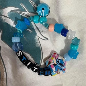 May include: A beaded bracelet featuring a variety of blue, light blue, and white plastic beads in star, round, and cube shapes. The bracelet includes black cube beads spelling out the word STITCH and a large star-shaped bead with a Stitch graphic.