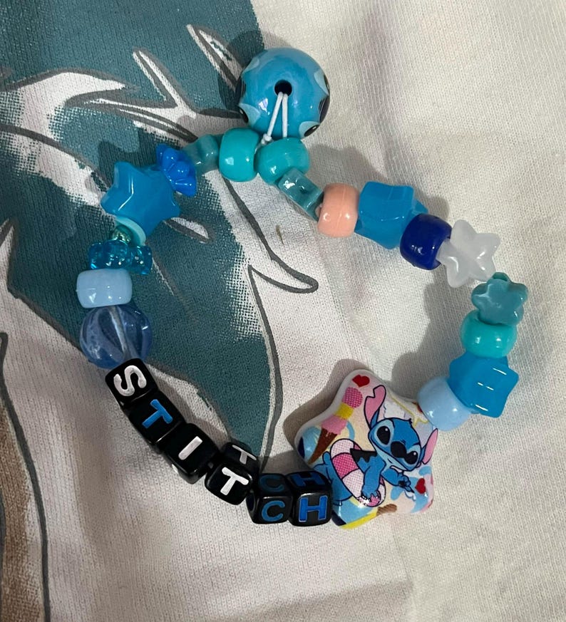 May include: A beaded bracelet featuring a variety of blue, light blue, and white plastic beads in star, round, and cube shapes. The bracelet includes black cube beads spelling out the word STITCH and a large star-shaped bead with a Stitch graphic.