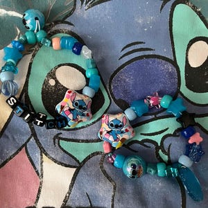 May include: Two beaded bracelets featuring blue, light blue, and pink plastic beads in various shapes including stars and spheres. Each bracelet includes a star-shaped charm depicting a blue character and black letter beads spelling STITCH.
