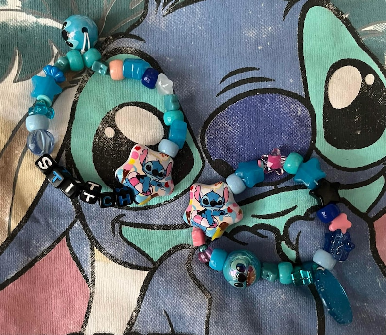 May include: Two beaded bracelets featuring blue, light blue, and pink plastic beads in various shapes including stars and spheres. Each bracelet includes a star-shaped charm depicting a blue character and black letter beads spelling STITCH.