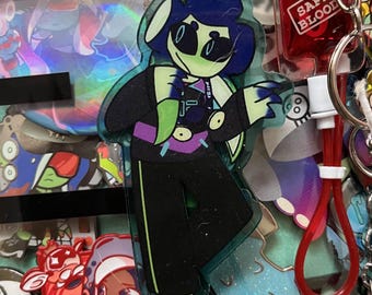 May include: A green, blue, and black cartoon character with a white headband and sunglasses. The character is wearing a black jacket with a green and purple design. The character is holding a green leaf in their right hand. The character is a keychain.