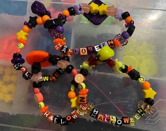 May include: Three Halloween-themed beaded bracelets. The bracelets feature orange, black, purple, and yellow beads, with star, smiley face, and fruit charms. The word "HALLOWEEN" is spelled out on some of the beads.