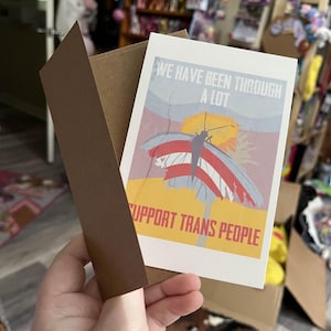 Puede incluir: Una tarjeta de felicitación con un sobre marrón. La tarjeta presenta una mariposa con alas rojas, blancas y azules, y el texto "WE HAVE BEEN THROUGH A LOT" y "SUPPORT TRANS PEOPLE". El fondo es una mezcla de amarillo, azul y rojo.