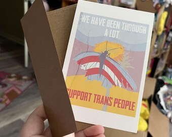 May include: A greeting card with a brown envelope. The card features a butterfly with red, white, and blue wings, and the text "WE HAVE BEEN THROUGH A LOT" and "SUPPORT TRANS PEOPLE". The background is a mix of yellow, blue, and red.