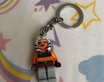 May include: A keychain featuring a Lego figure of Ahsoka Tano from Star Wars. The figure has orange skin, blue and white markings, and gray pants. The keychain has a silver chain and ring.