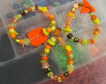 May include: A collection of colorful beaded bracelets with a pumpkin theme. The bracelets feature orange, yellow, and clear beads, with some spelling out "PUMPKIN". Other beads include stars, smiley faces, and orange shapes. The bracelets are arranged in a clear plastic container.
