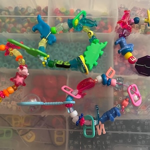 Könnte beinhalten: Mehrere bunte Armbänder mit Perlen und verschiedenen Anhängern, darunter Sterne, Tiere und geometrische Formen. Die Armbänder befinden sich in einem transparenten Kunststoffbehälter, der eine Vielzahl von Farben und Designs zeigt. Die Perlen sind aus Kunststoff und die Anhänger aus Kunststoff und Metall.