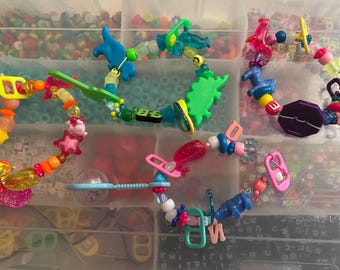 May include: Several colorful beaded bracelets with various charms, including stars, animals, and geometric shapes. The bracelets are in a clear plastic container, showcasing a variety of colors and designs. The beads are made of plastic and the charms are made of plastic and metal.