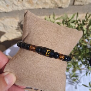 May include: A stretch bracelet featuring smooth black beads and brown tiger eye beads. A central cylindrical black bead displays the letter E in gold. The beads are arranged in a repeating pattern along the elastic cord of the jewelry piece.