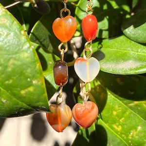 May include: Gold heart-shaped earrings with carnelian and agate beads. The earrings have a layered design with three beads on each earring.
