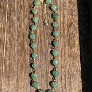 Pu&ograve; includere: Una collana con perline verdi a forma di cuore collegate da una catena color oro. La collana ha una chiusura color oro e un piccolo ciondolo a forma di cuore all'estremit&agrave; della catena regolabile. La collana &egrave; esposta su una superficie di legno.