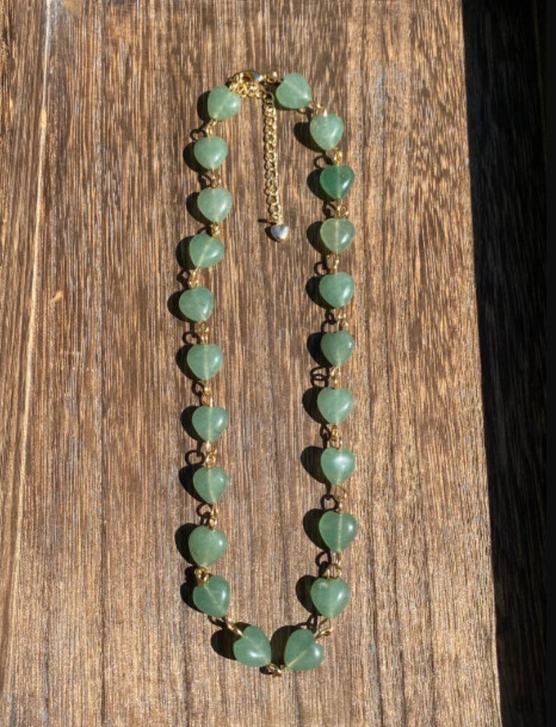 Pu&ograve; includere: Una collana con perline verdi a forma di cuore collegate da una catena color oro. La collana ha una chiusura color oro e un piccolo ciondolo a forma di cuore all'estremit&agrave; della catena regolabile. La collana &egrave; esposta su una superficie di legno.