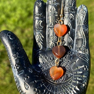 May include: A pair of gold heart-shaped earrings with three carnelian stones. The stones are orange, brown, and orange. The earrings are hanging from a black hand.