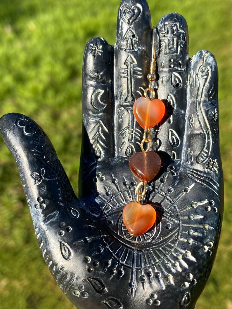 May include: A pair of gold heart-shaped earrings with three carnelian stones. The stones are orange, brown, and orange. The earrings are hanging from a black hand.