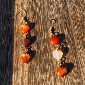 May include: A pair of gold dangle earrings with three heart-shaped carnelian stones on each earring. The stones are in shades of orange, brown, and white.