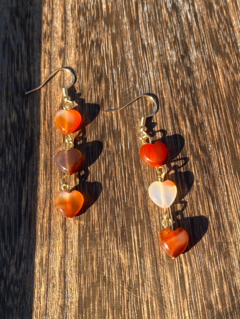 May include: A pair of gold dangle earrings with three heart-shaped carnelian stones on each earring. The stones are in shades of orange, brown, and white.