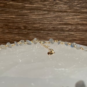 Puede incluir: Un collar de cadena dorada con un dije de coraz&oacute;n y una fila de piedras azul claro. El collar est&aacute; sobre una superficie blanca.