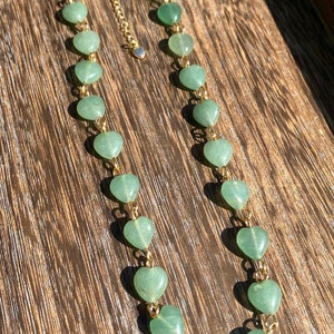 Pu&ograve; includere: Una delicata collana con pietre preziose verdi a forma di cuore infilate su una catena color oro. La collana ha un piccolo ciondolo a forma di cuore sulla chiusura, esposta su una superficie di legno.